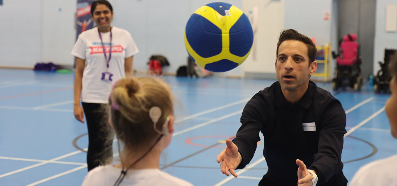 A girl wearing a hearing device passes a ball with a coach on an indoors court under the supervision a woman holding a folder.
