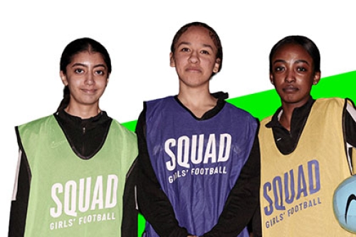 Three girls stand side-by-side wearing bibs with the words "SQUAD GIRLS' FOOTBALL" on them. One of the girls is also carrying a football under her left arm.